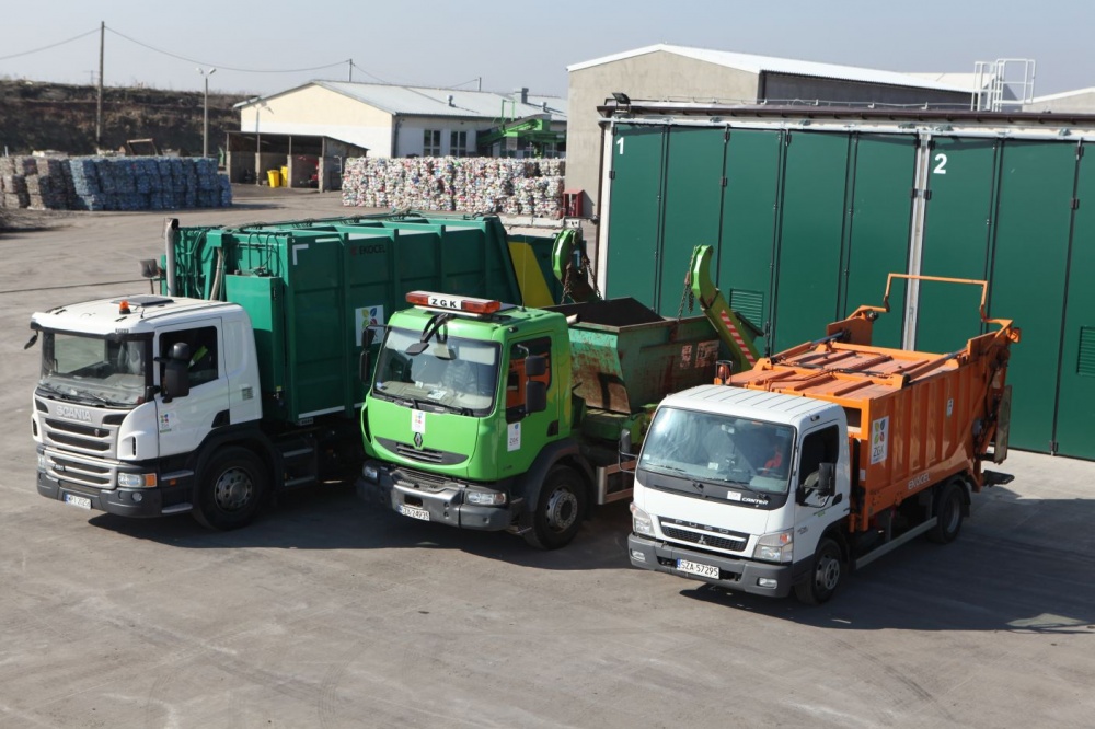 3 samochody ciężarowe- biało-zielona śmieciarka, zielony kontenerowiec oraz biało- pomarańczowy kontenerowiec