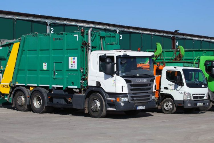 3 samochody ciężarowe- biało-zielona śmieciarka, zielony kontenerowiec oraz biało- pomarańczowy kontenerowiec